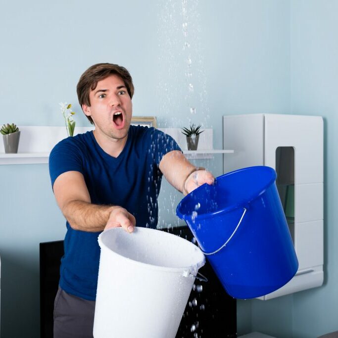 Young Male Worried Man Holding Bucket While Water Droplets Leaking From Ceiling